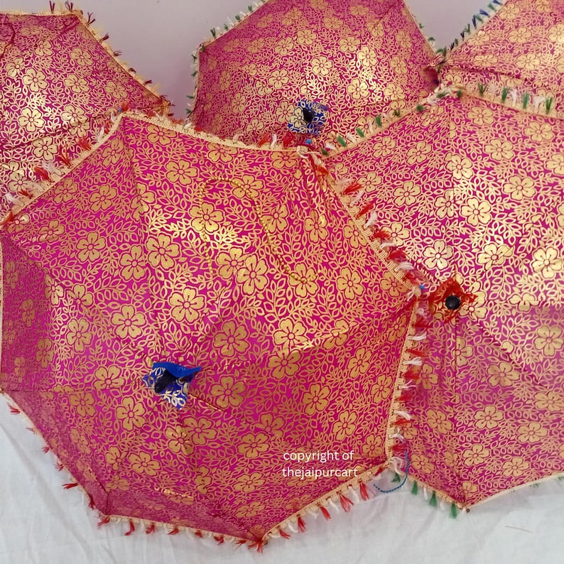 Puede incluir: Tres paraguas rosas con patrones florales dorados y ribetes de flecos. Los paraguas est&aacute;n plegados y apilados uno encima del otro. El texto "copyright of thejaipurbcart" es visible en la esquina inferior derecha.