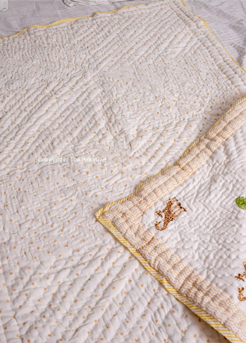 K&ouml;nnte beinhalten: Wei&szlig;e und gelbe Babydecke mit einem kleinen Giraffenmuster. Die Decke ist mit einem Rautenmuster gesteppt und hat einen gelben Rand.