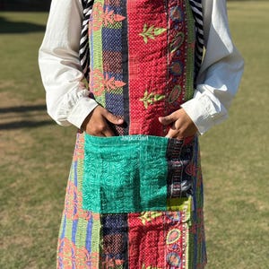 May include: A patchwork apron with a large front pocket. The apron features a variety of colorful fabrics, including red, green, blue, and patterned designs. The apron is quilted and has a neck strap and waist ties.
