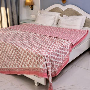 May include: A pink and white cotton quilt with a paisley print. The quilt is folded on a white bed frame with white pillows.