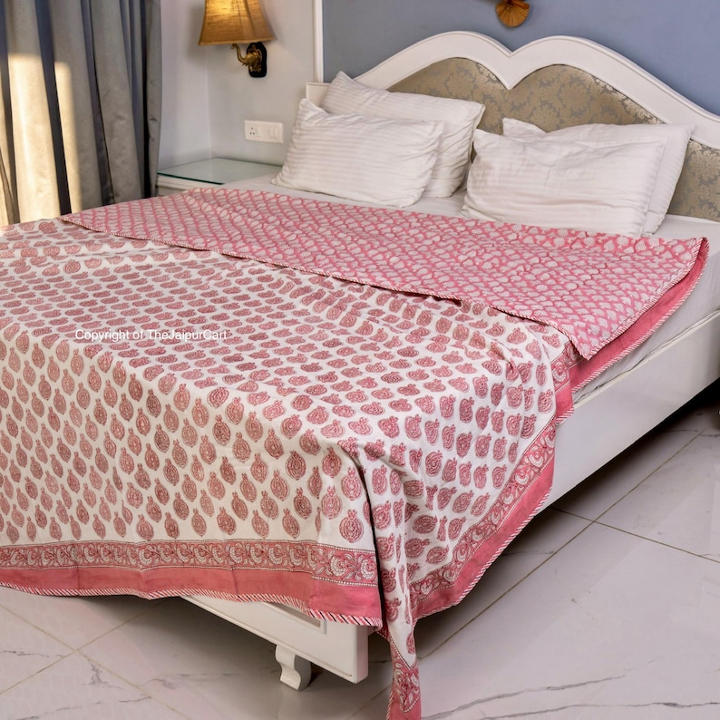 May include: A pink and white cotton quilt with a paisley print. The quilt is folded on a white bed frame with white pillows.