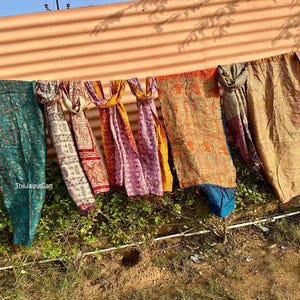 May include: A collection of colorful, patterned scarves and textiles hanging on a clothesline. The textiles feature various designs and colors, including teal, cream, red, pink, orange, and gold. The items are displayed outdoors.