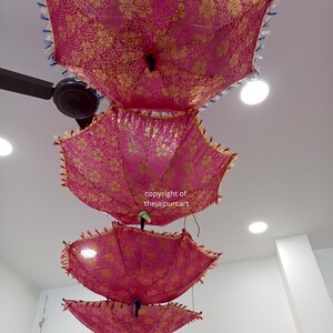Puede incluir: Tres paraguas rosas y dorados con patrones florales cuelgan del techo. Los paraguas est&aacute;n superpuestos, siendo el paraguas superior el m&aacute;s grande y el inferior el m&aacute;s peque&ntilde;o. El texto "copyright of thejaipurcart" es visible en la imagen.