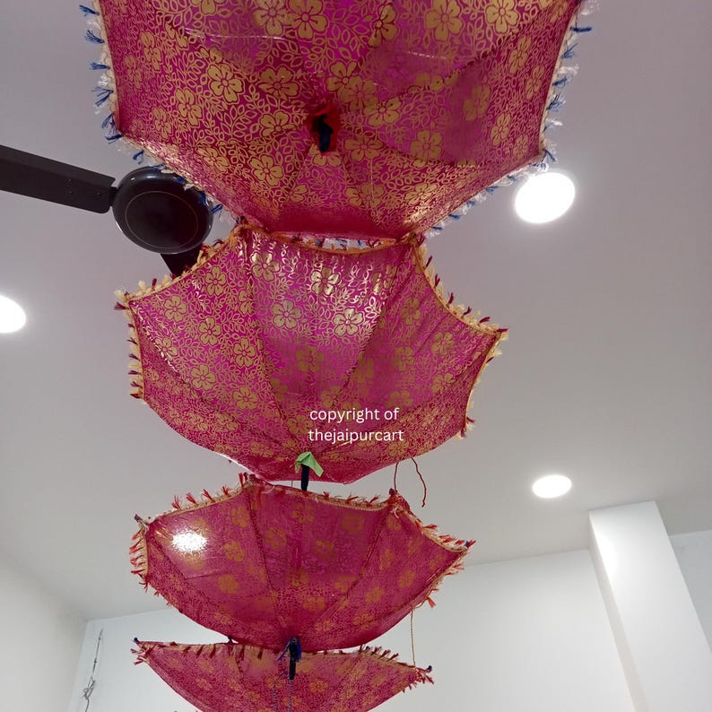 Puede incluir: Tres paraguas rosas y dorados con patrones florales cuelgan del techo. Los paraguas est&aacute;n superpuestos, siendo el paraguas superior el m&aacute;s grande y el inferior el m&aacute;s peque&ntilde;o. El texto "copyright of thejaipurcart" es visible en la imagen.