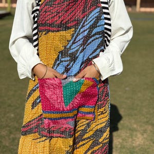 May include: A colorful, patchwork apron with a large front pocket. The apron features a vibrant mix of red, blue, yellow, and pink fabrics, stitched together with visible thread. The apron is designed with a neck strap and waist ties.