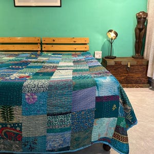 Puede incluir: Un edredón de retazos en tonos turquesa, azul y verde cubre una cama. El edredón presenta una variedad de telas estampadas, cosidas juntas. La cama tiene un cabecero de madera. Una obra de arte enmarcada cuelga de la pared sobre la cama.