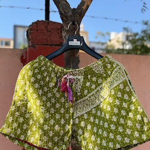 May include: Green shorts with a white floral design and red trim, hanging on a black hanger. The shorts feature a drawstring with a tassel. The shorts are made from quilted fabric. The shorts are approximately 30 cm in length.