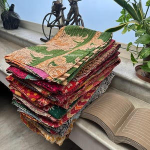 以下が含まれることがあります： リサイクルされたサリーから作られたカラフルなカンタキルトの積み重ね。緑とオレンジのキルトが上にあります。キルトは折り畳んで白い表面に積み重ねられています。緑とオレンジのキルトに「TheJaipurCart」という文字が見えます。