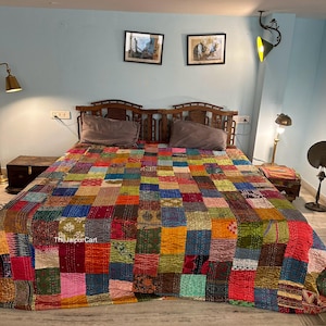 May include: A colorful patchwork quilt made from recycled saris, featuring a variety of colors and patterns. The quilt is laid out on a bed with a wooden headboard and two pillows. The quilt is made in India and is available for purchase on TheJaipurCart.