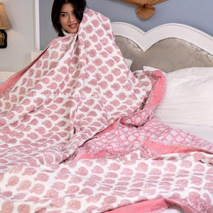 May include: A pink and white patterned cotton quilt with a floral design. The quilt is folded on a white bed with a white pillow.