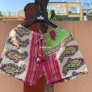 Op de afbeelding: Korte broek met een witte achtergrond en groene paisley-patroon, geaccentueerd met roze verticale strepen en een groen paneel. De broek hangt aan een zwarte hanger tegen een vage buitenachtergrond.