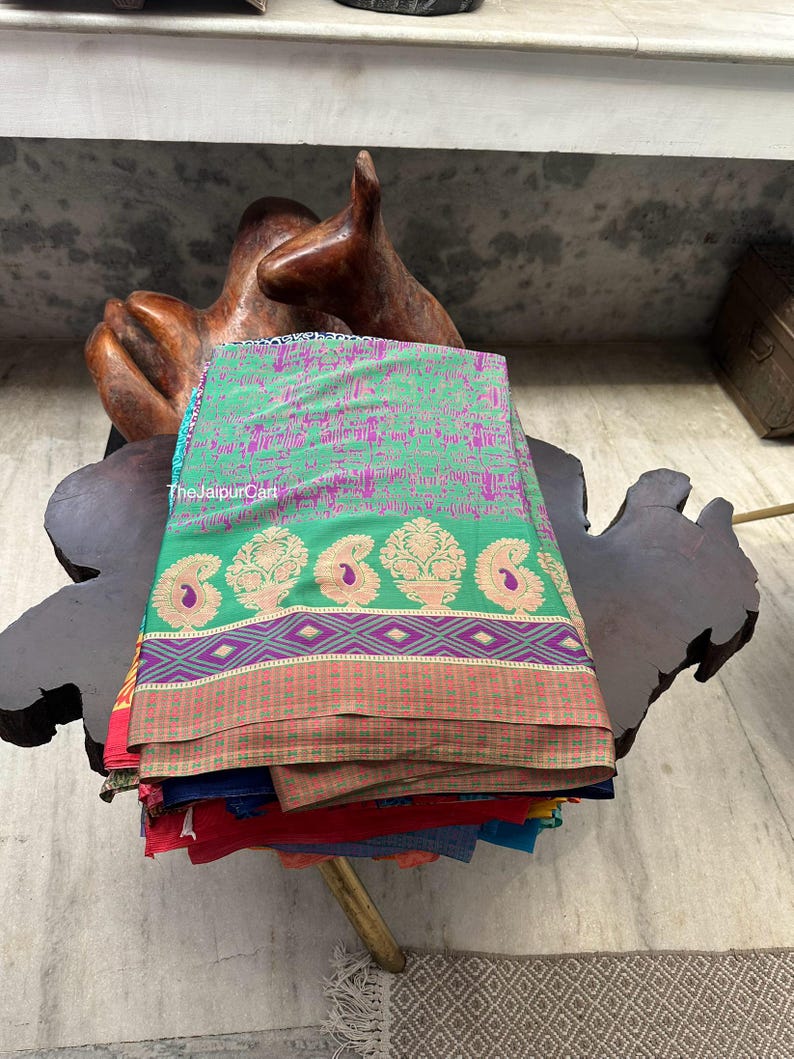 May include: A stack of colorful fabric saris with a green and gold paisley design. The saris are folded and stacked on a wooden table.