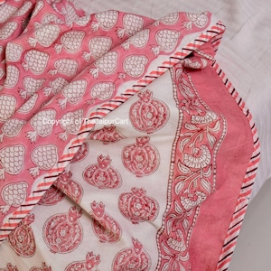 May include: A pink and white cotton quilt with a pomegranate print. The quilt has a white background with pink pomegranates and a pink border with a white floral design. The quilt is folded in half and the edges are visible.
