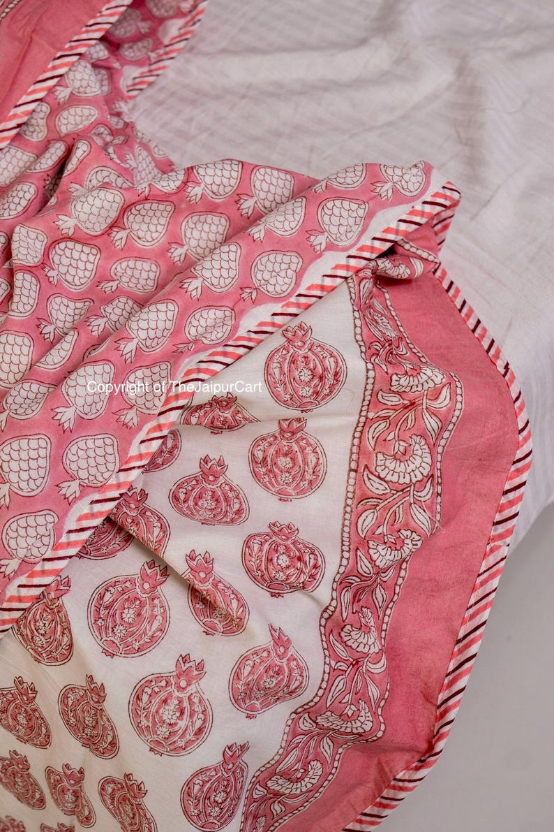 May include: A pink and white cotton quilt with a pomegranate print. The quilt has a white background with pink pomegranates and a pink border with a white floral design. The quilt is folded in half and the edges are visible.