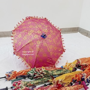 Puede incluir: Un paraguas rosa y dorado con un borla azul. El paraguas est&aacute; cerrado y tiene un borde de flecos. El paraguas est&aacute; rodeado de otros paraguas de varios colores y patrones.