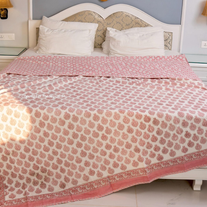May include: A white cotton quilt with a pink floral pattern. The quilt is folded on a bed with white pillowcases.