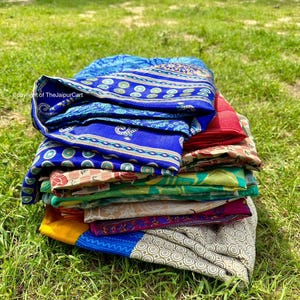 May include: A stack of colorful, folded textiles rests on green grass. The textiles display a variety of patterns and colors, including blue, red, yellow, and green. The textiles appear to be made of a lightweight fabric.