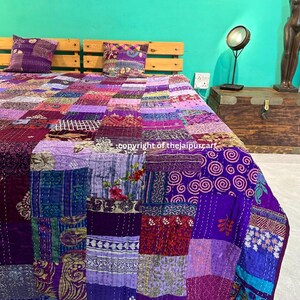 May include: A purple and blue patchwork quilt featuring a variety of floral and geometric patterns. The quilt is made from kantha fabric and is hand-stitched. The quilt is displayed on a bed with two pillows.