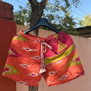May include: Orange shorts with a colorful geometric pattern in pink and green. The shorts are hanging on a black hanger. The shorts have a drawstring waist and a decorative tassel.