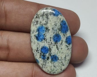 May include: An oval-shaped K2 stone, featuring a speckled white and gray base with vibrant blue orb-like patterns. The polished gemstone is held in a hand, showcasing its natural beauty and unique color variations.