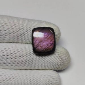 Könnte beinhalten: Ein quadratischer Labradorit-Edelstein mit abgerundeten Ecken, der ein leuchtendes violettes und magentafarbenes Schillern auf seiner Oberfläche zeigt, gekennzeichnet durch ein Netzwerk feiner, dunkler Risse und eine glatte Politur.