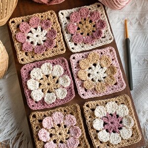 May include: Six handmade crochet squares in shades of pink, beige, and white, arranged on a wooden surface. Each square features a floral design in the center. Yarn balls and a crochet hook are visible in the background, suggesting a crafting theme.