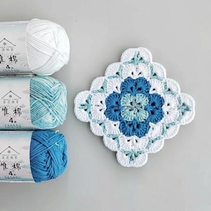 May include: A crocheted square coaster in white, light blue, and dark blue. Beside it are three skeins of yarn in matching colours. The yarn skeins have Chinese text on the label.