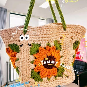 Op de afbeelding: Een beige gehaakte tote bag met een groot zonnebloemontwerp in oranje en groen. De tas heeft groene handvatten en de tekst "Yummy Food" is zichtbaar. De tas is handgemaakt met een granny square patroon.