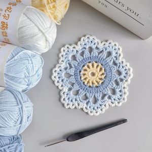 Puede incluir: Un posavasos de ganchillo en tonos azules, blancos y amarillos, con un diseño floral. Se ven varios ovillos de lana en colores a juego, junto con un ganchillo. El texto del embalaje del hilo es borroso.