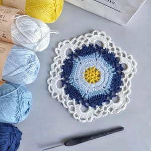 Könnte beinhalten: Ein handgefertigter Häkeluntersetzer mit einem Blumendesign in Blau-, Weiß- und Gelbtönen. Der Untersetzer ist von weißem, blauem und gelbem Garn sowie einer Häkelnadel umgeben.