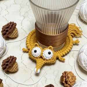May include: A handmade yellow crochet coaster with a whimsical worm design, featuring large white-rimmed eyes and a white nose. A clear glass with a wooden sleeve sits atop the coaster. Decorative crochet pinecone-shaped items and white yarn balls are scattered around.