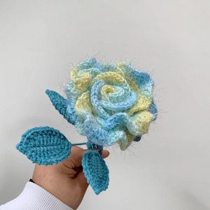 Pode incluir: Uma rosa de crochê feita à mão em tons de azul e amarelo. A flor tem um design espiral detalhado, com três folhas turquesas. O caule é azul escuro. A flor é segurada por uma pessoa.