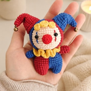 Peut inclure: Une petite poupée clown au crochet avec un chapeau de bouffon rouge et bleu orné de minuscules clochettes métalliques aux extrémités. La poupée présente un visage crème, un nez rouge vif, des yeux en perles noires et une collerette jaune.