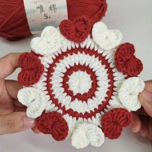 May include: A handmade crochet coaster with a heart design. The coaster features alternating red and white hearts around the edge and a concentric circle pattern in the center. A ball of red yarn is in the background.