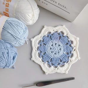 May include: A handmade crochet coaster with a floral design in white and blue hues. The coaster is next to balls of white and light blue yarn, a crochet hook, and a book with the text "No one can replace you".