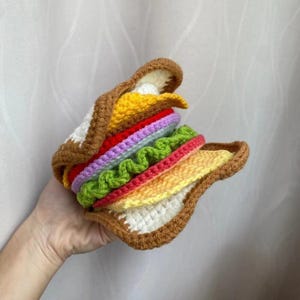 Peut inclure: Un sandwich crocheté fait à la main, avec des couches de garnitures jaunes, rouges, vertes et violettes entre deux tranches de pain marron et blanc. Le sandwich est tenu dans une main, mettant en valeur le travail de crochet détaillé et les couleurs vives.