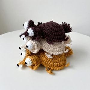 Puede incluir: Una pila de cuatro figuras de animales de ganchillo en tonos marrón, beige y amarillo. Cada figura tiene grandes ojos blancos con pupilas negras y pequeñas colas rizadas. Las figuras están apiladas unas sobre otras.