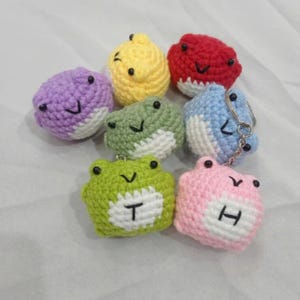 Puede incluir: Una colección de llaveros de rana hechos a mano con ganchillo en varios colores, incluyendo verde, rosa, morado, amarillo, rojo y azul. Cada rana tiene una barriga blanca y ojos negros, y algunas presentan las letras "T" y "H".