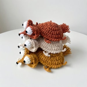 May include: A stack of four crocheted animal figures in shades of brown, tan, and yellow. Each figure has large white eyes with black pupils and small, detailed legs. The figures are stacked on top of each other.