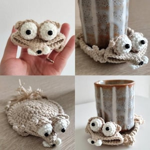 May include: Crocheted beige and white cup coasters in the shape of a cartoon animal with large eyes. The coasters are shown with a ceramic mug and are handmade. The animal has large, round eyes and a small nose.