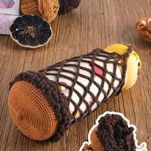 May include: A handmade, brown crochet pencil case with a yellow and white object inside. The case has a net-like design and is decorated with a brown crochet border. A small, brown crochet decoration is also visible.