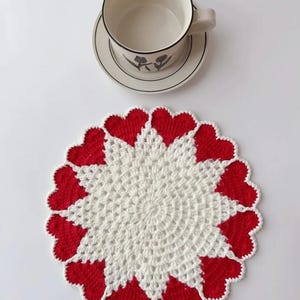 May include: A white and red crocheted coaster with a heart-shaped border. A white teacup and saucer with a black floral design are on a white surface. The coaster is handmade and adds a decorative touch to any table setting.