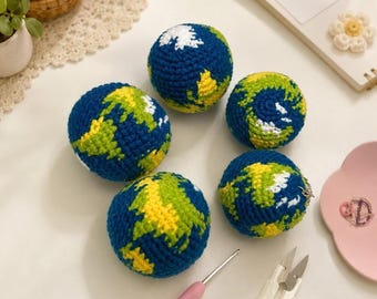 May include: Five handmade crochet balls depicting the Earth. The balls are primarily blue, with green, yellow, and white sections representing continents and ice caps. A pink crochet hook and small scissors are also visible.