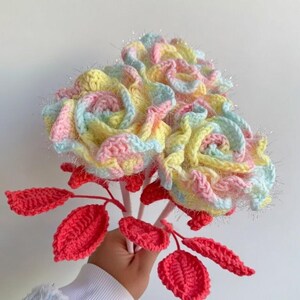 May include: A bouquet of handmade crochet flowers in pastel colors, including yellow, pink, and blue. The flowers have a textured appearance with silver accents. Red crochet leaves and pink stems complete the floral arrangement.