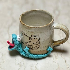 Può includere: Una tazza di ceramica maculata con una lucertola blu all'uncinetto avvolta attorno. La tazza ha un'illustrazione in bianco e nero di un gatto. La lucertola ha una lingua rossa e occhi bianchi.