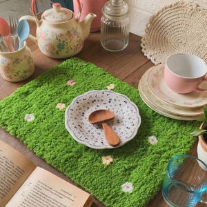 Puede incluir: Naturaleza muerta con un mantel individual verde con forma de hierba adornado con pequeños detalles florales. Una tetera con estampado floral, platos y una taza rosa están dispuestos sobre una superficie de madera, junto con una cuchara de madera y un libro.