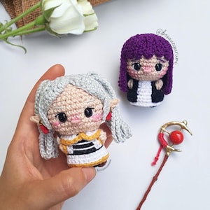 Puede incluir: Muñecas de ganchillo con cabello gris y morado, vestidas con trajes blancos y negros. Una muñeca se sostiene en una mano. También hay un bastón rojo y dorado. Las muñecas tienen mejillas rosadas y ojos grandes. Fondo blanco con rosas blancas.