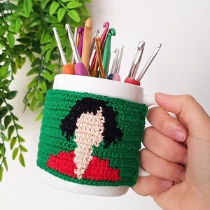 May include: A white mug with a green crocheted cozy featuring a woman's portrait in red, black, and beige. The mug holds various crochet hooks. The text "@BOSQUEGURUMI" is visible in the upper right corner.