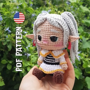 Puede incluir: Una muñeca de ganchillo con cabello gris, orejas puntiagudas y un vestido blanco con ribetes dorados y rayas negras. La muñeca tiene mejillas rosadas y ojos negros grandes. Incluye el texto "PDF PATTERN" y la marca "Bosquegurumi".