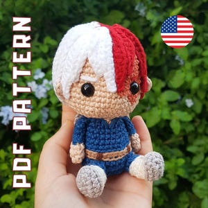 Puede incluir: Muñeca amigurumi de crochet con pelo blanco y rojo, ropa azul y cara beige. La muñeca está siendo sostenida en una mano.